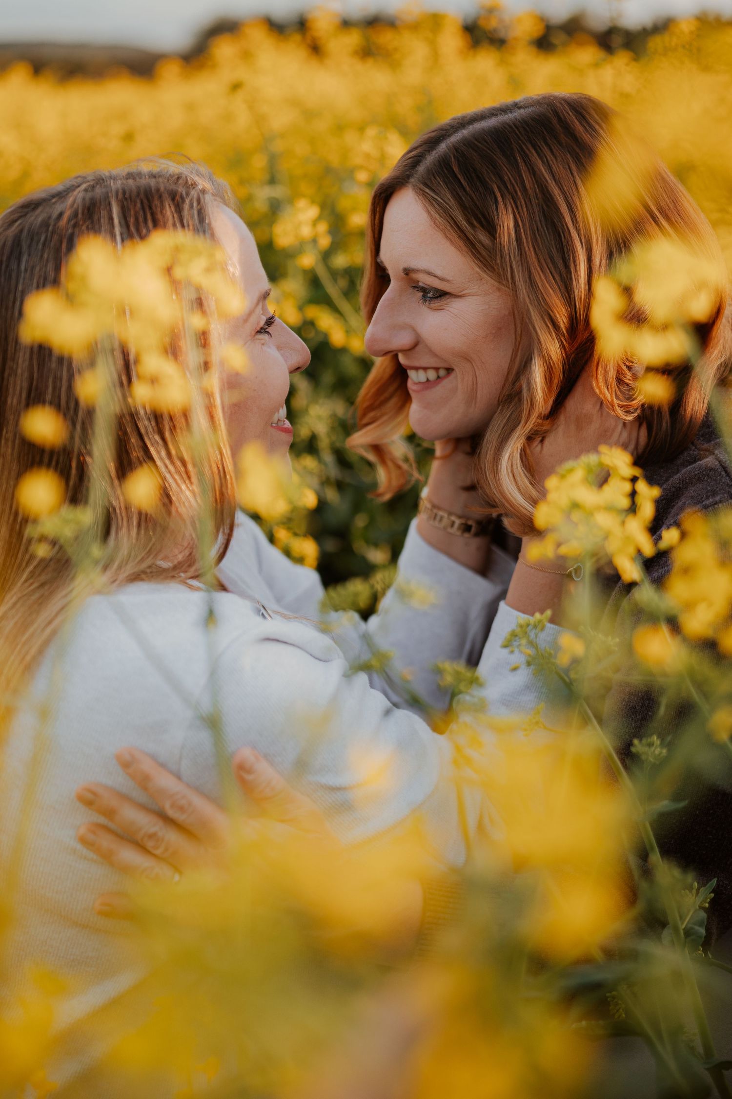 Mother and daughter smiling at each other in a vibrant yellow rapeseed flower field during golden hour.