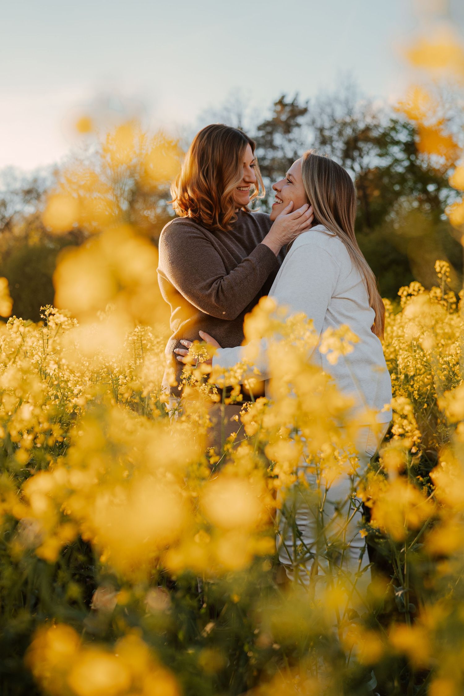Two women embrace and smile at each other in a vibrant yellow wildflower field during golden hour.