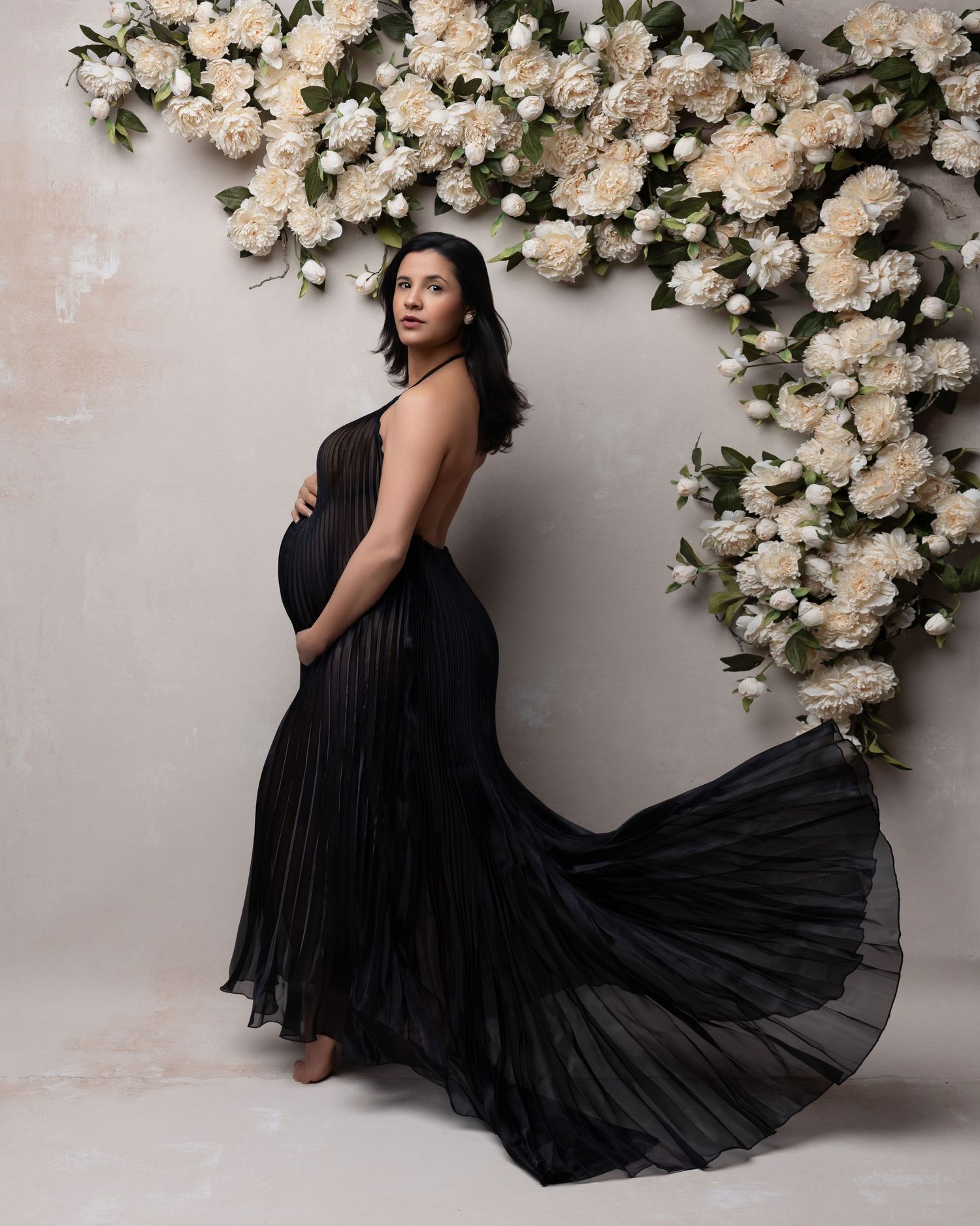 Elegant black pleated dress flows gracefully against white floral backdrop in romantic studio photoshoot.