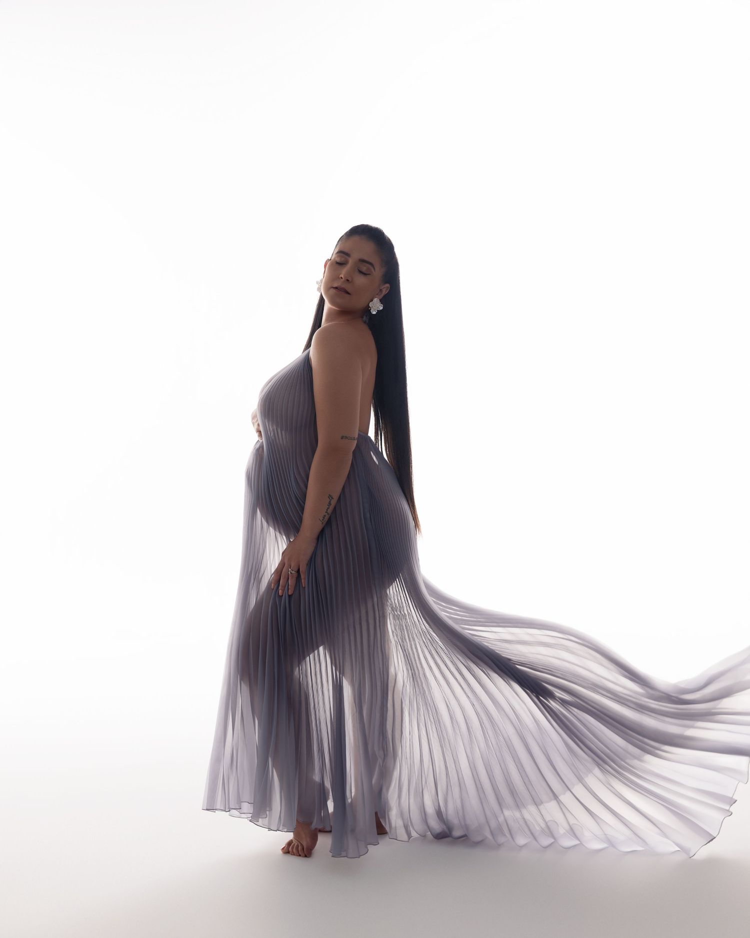 A person poses gracefully in a flowing pleated lavender gown against a white background, creating an ethereal effect.