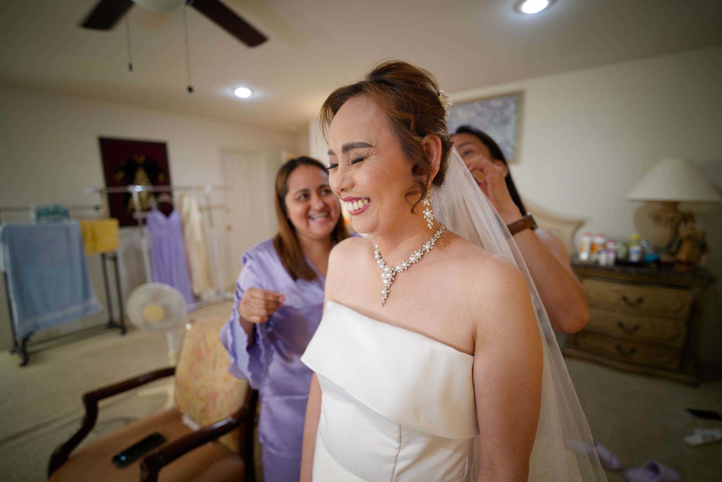 A glowing bride wearing a white strapless dress and crystal necklace smiles while getting ready for her wedding day.