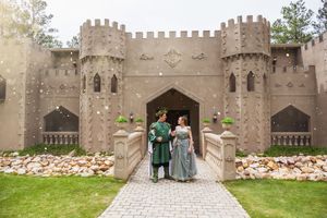 Two figures in medieval-style costumes pose in front of an ornate castle facade with stone towers and crenellations.