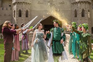 A whimsical medieval reenactment scene shows performers in period costumes dancing in a line outside a stone castle.