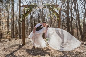 A romantic wedding scene under a rustic wooden arch in a sun-dappled forest setting with flowing white veil catching the breeze.