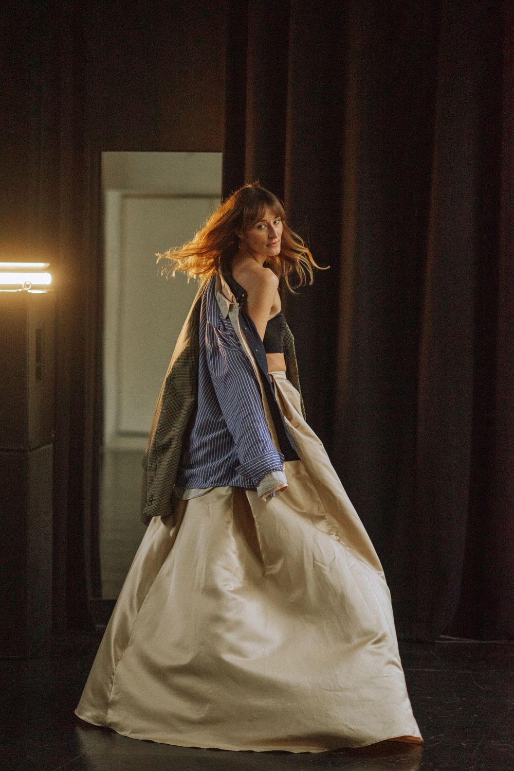 Dancer in white dress and denim jacket spins gracefully against dark curtains.
