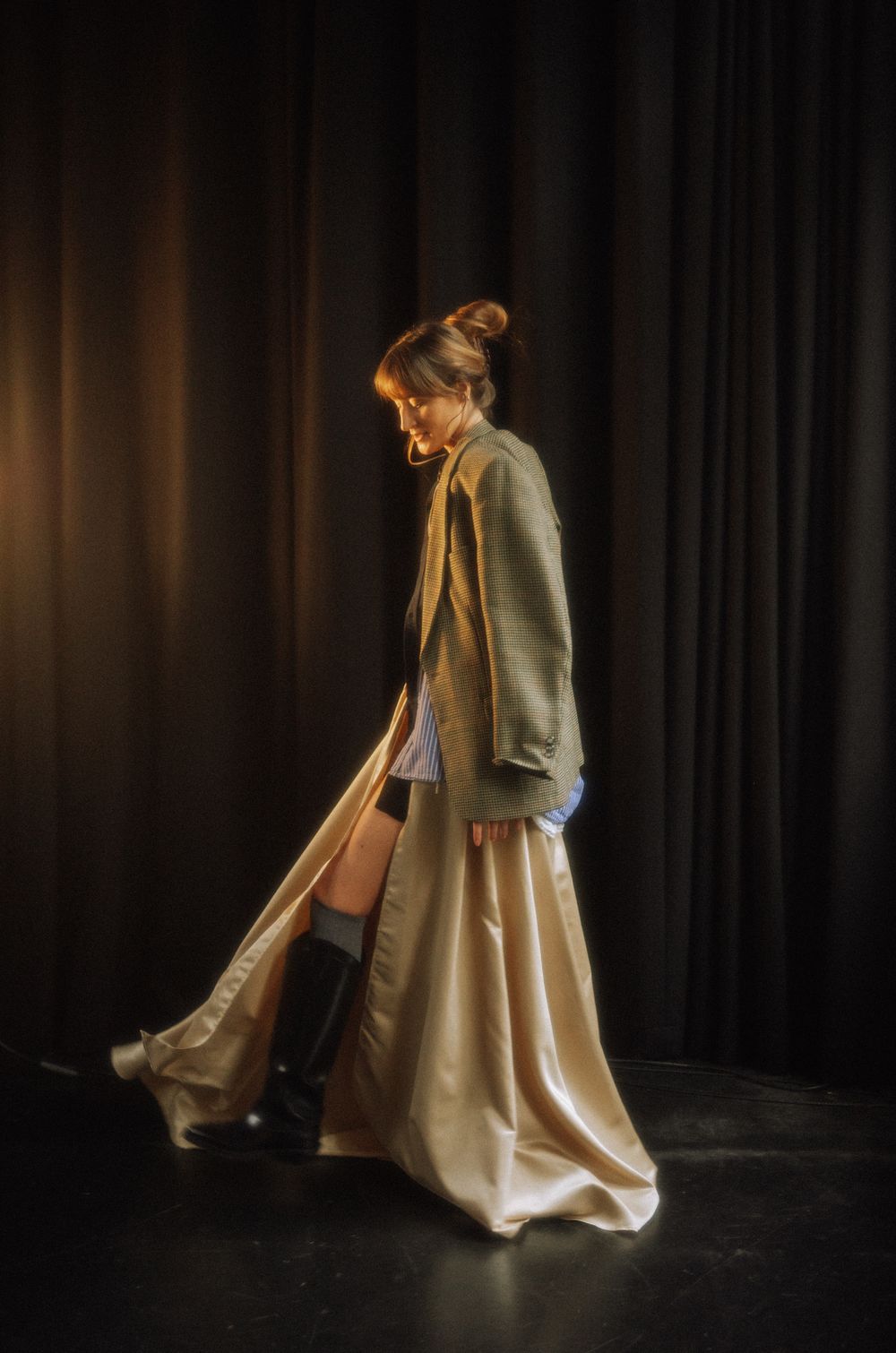 Model wearing flowing beige dress and blazer in theatrical lighting against dark curtains.