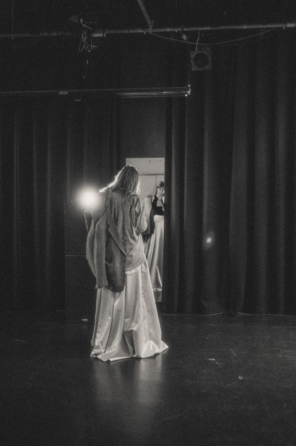 Silhouette in flowing dress standing on dark stage with theatrical lighting.