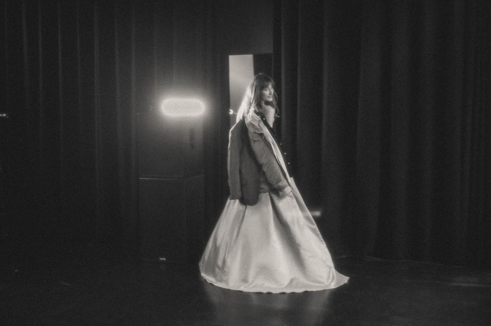 Black and white stage photo of figure in flowing dress illuminated by spotlight.