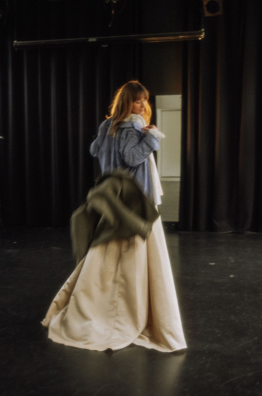 A figure in flowing cream fabric moves dramatically against a dark backdrop on stage.
