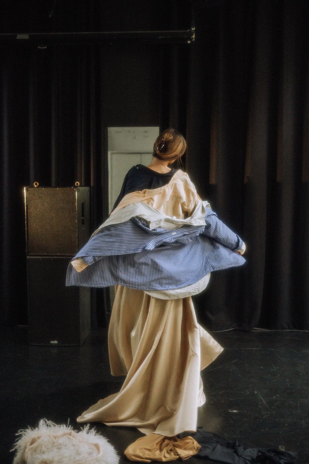 Dancer in cream dress removes blue striped fabric while performing on dark stage.