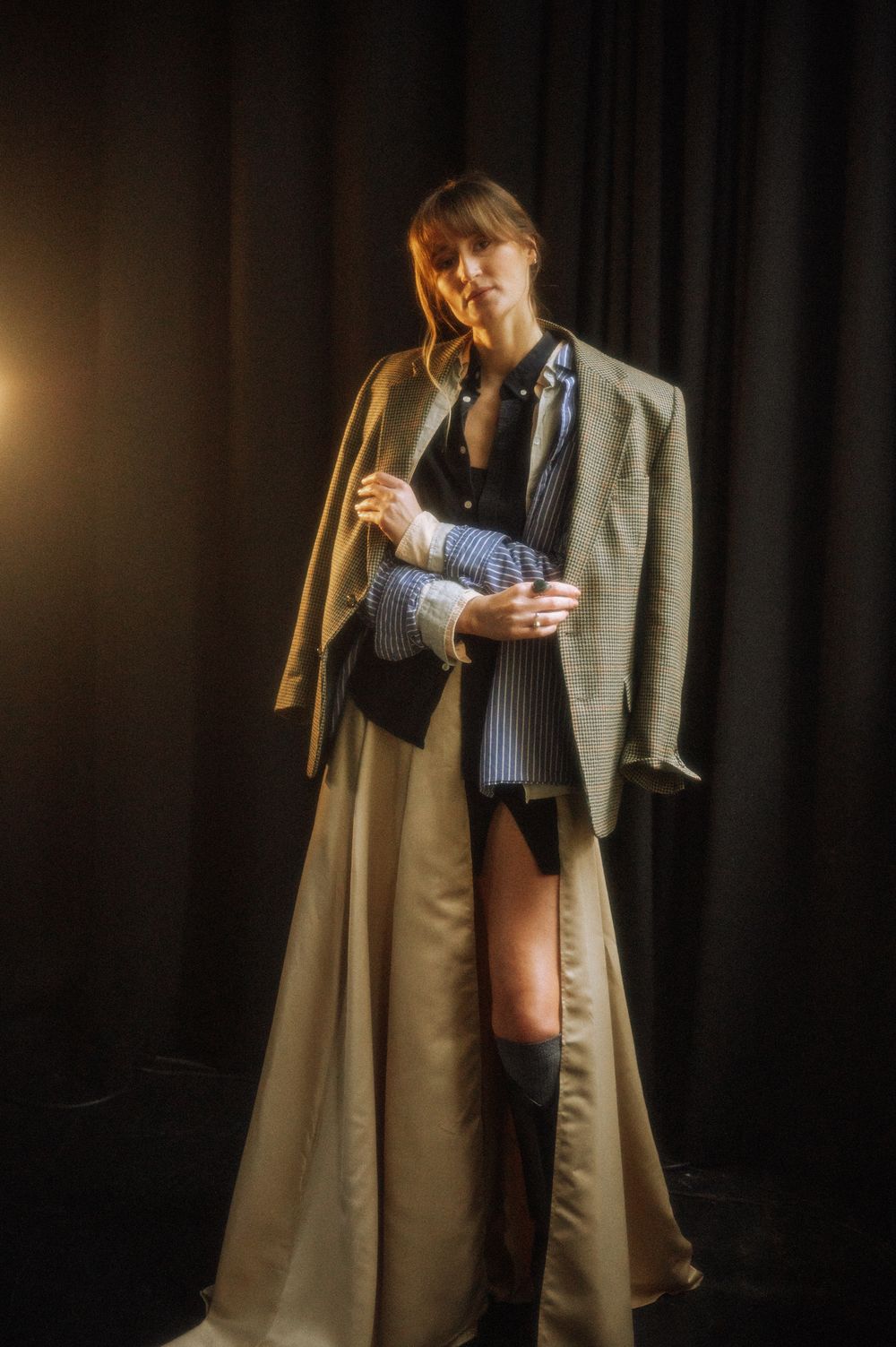 Dramatic fashion photo showing long beige coat and layered styling against dark backdrop.
