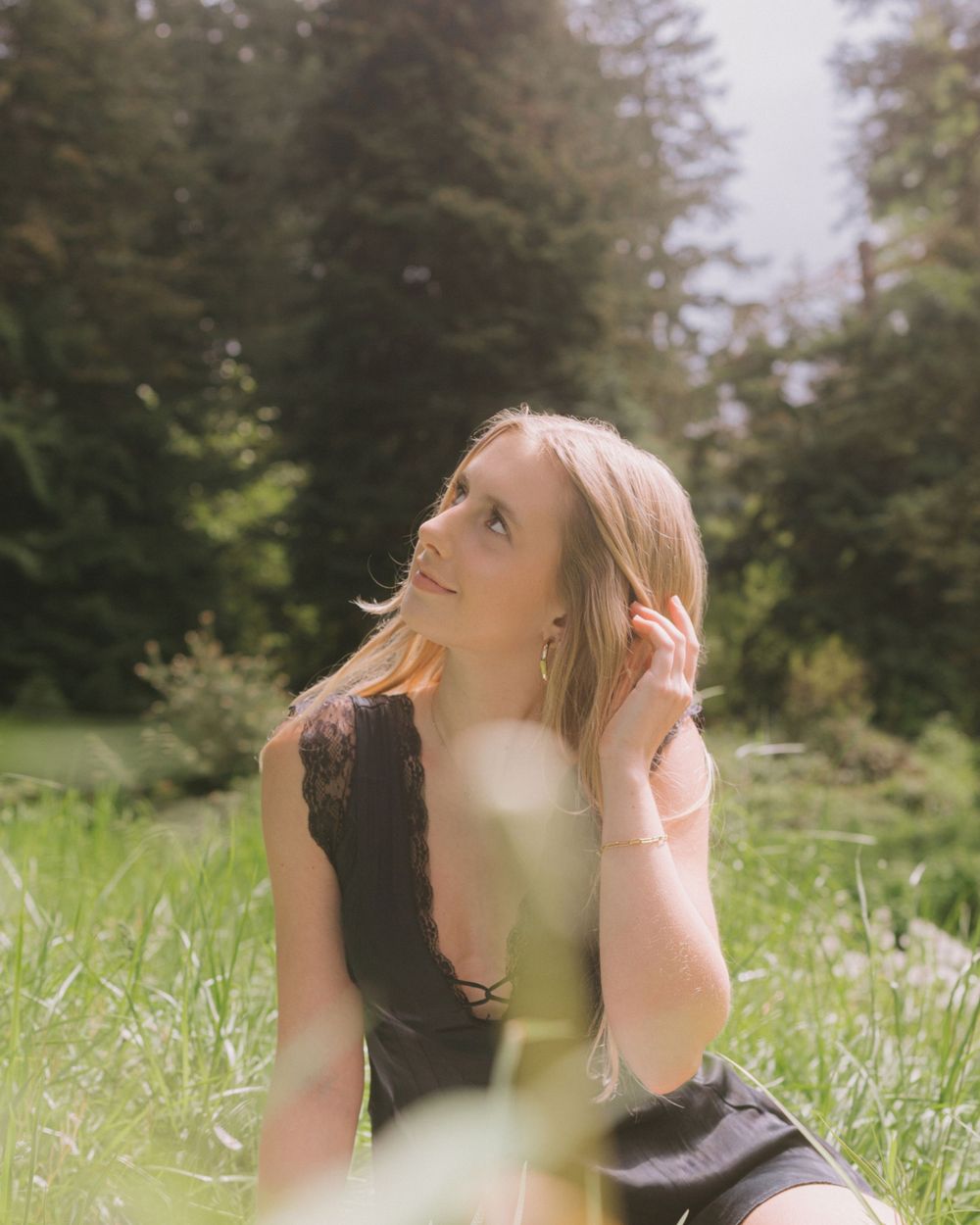 High school senior in dark dress poses thoughtfully in sunlit meadow with trees.