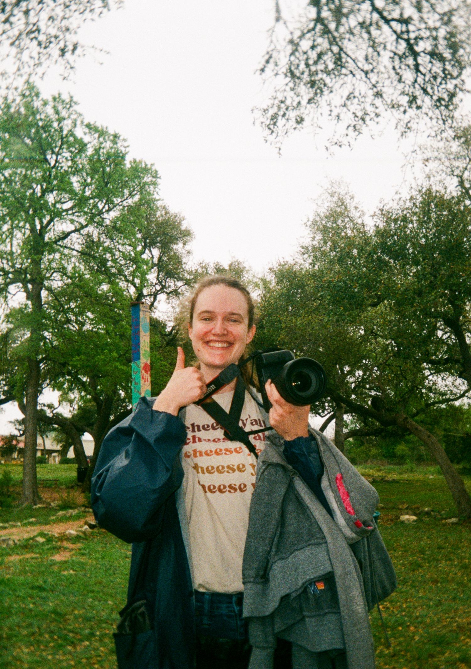 Wedding and portrait photographer holding camera and making thumbs up sign while standing in green outdoor setting.