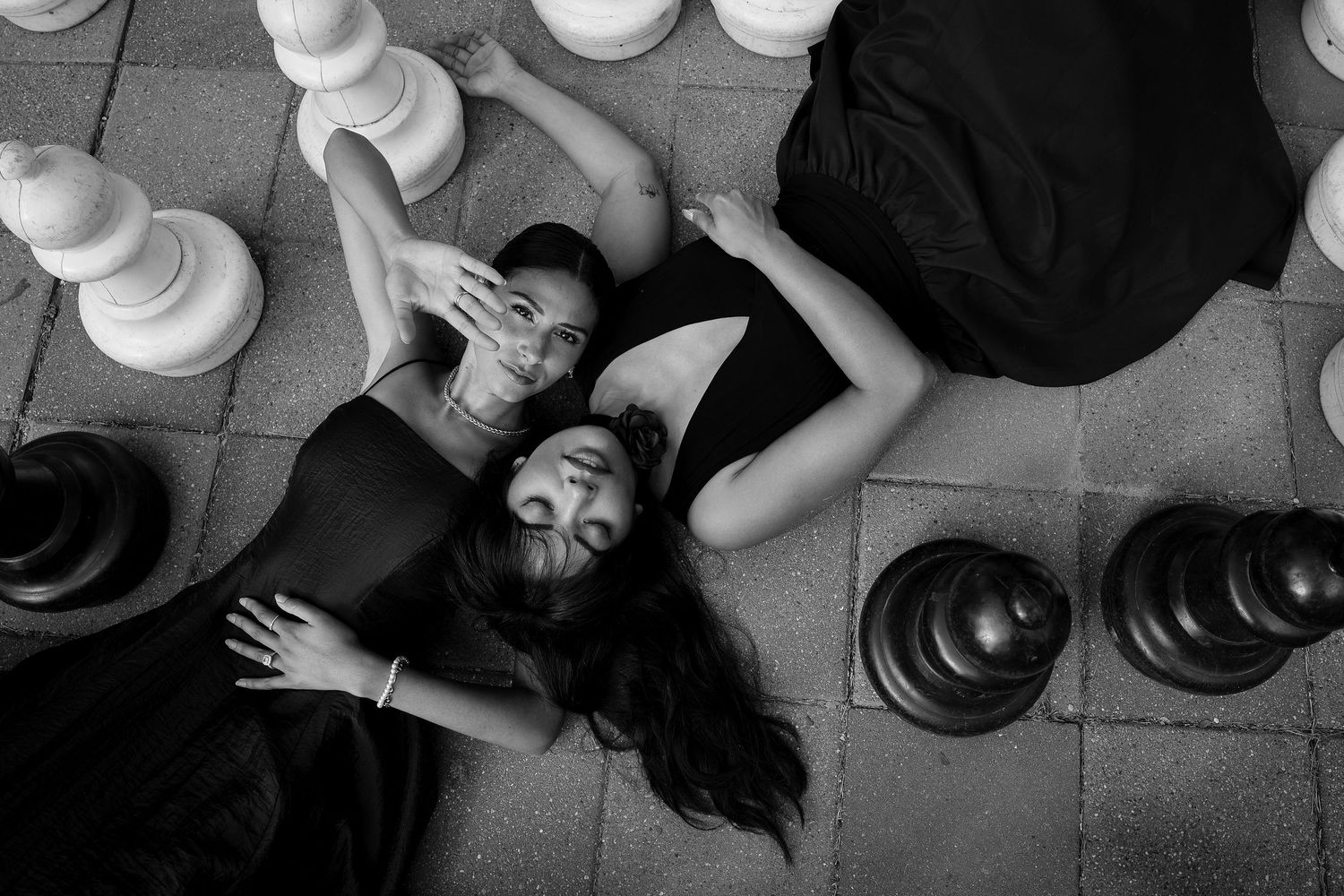 An artistic black and white photo of people lying on floor surrounded by chess pieces.