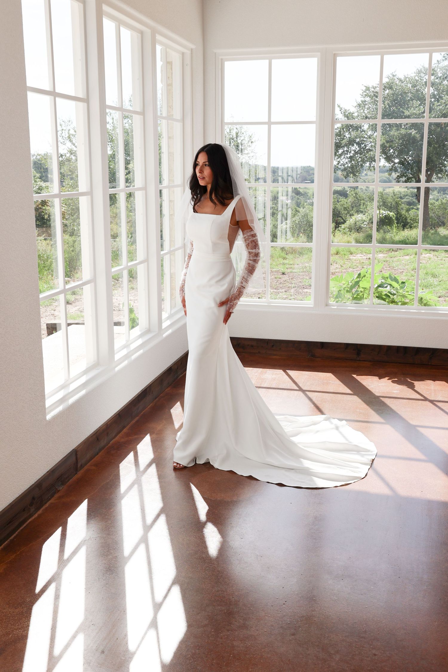 A bride in a fitted white long-sleeve wedding dress poses by sunlit windows in an elegant room.
