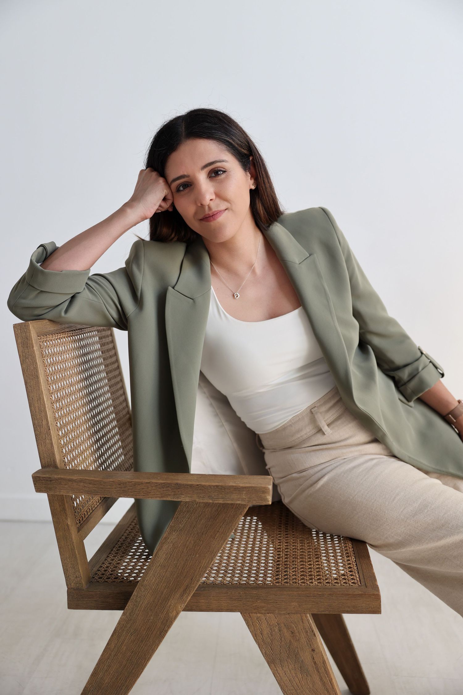 A model in a sage green blazer and cream outfit poses on a wooden cane chair.