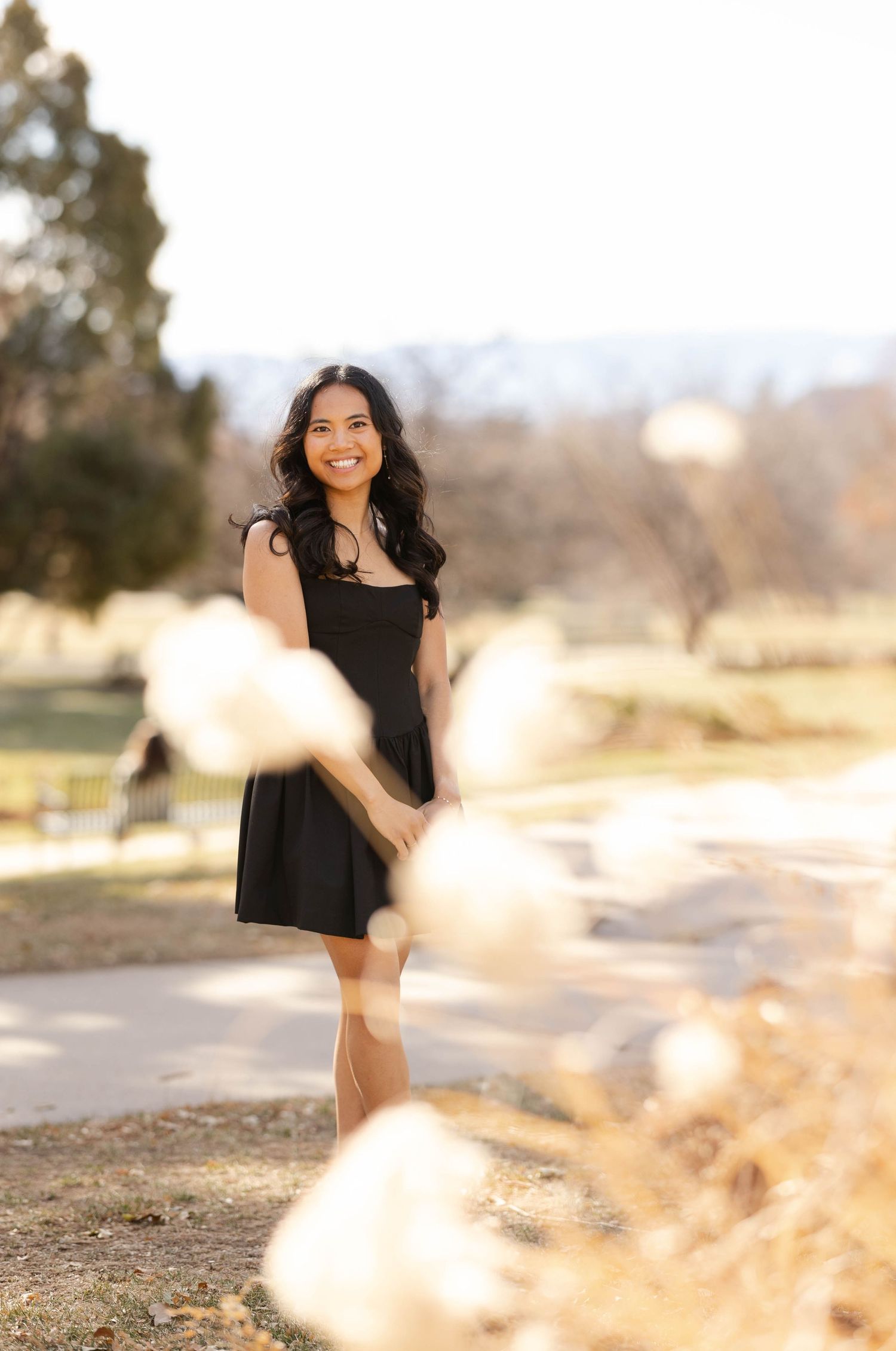 A woman in a black dress poses outdoors with blurred natural elements in the foreground.