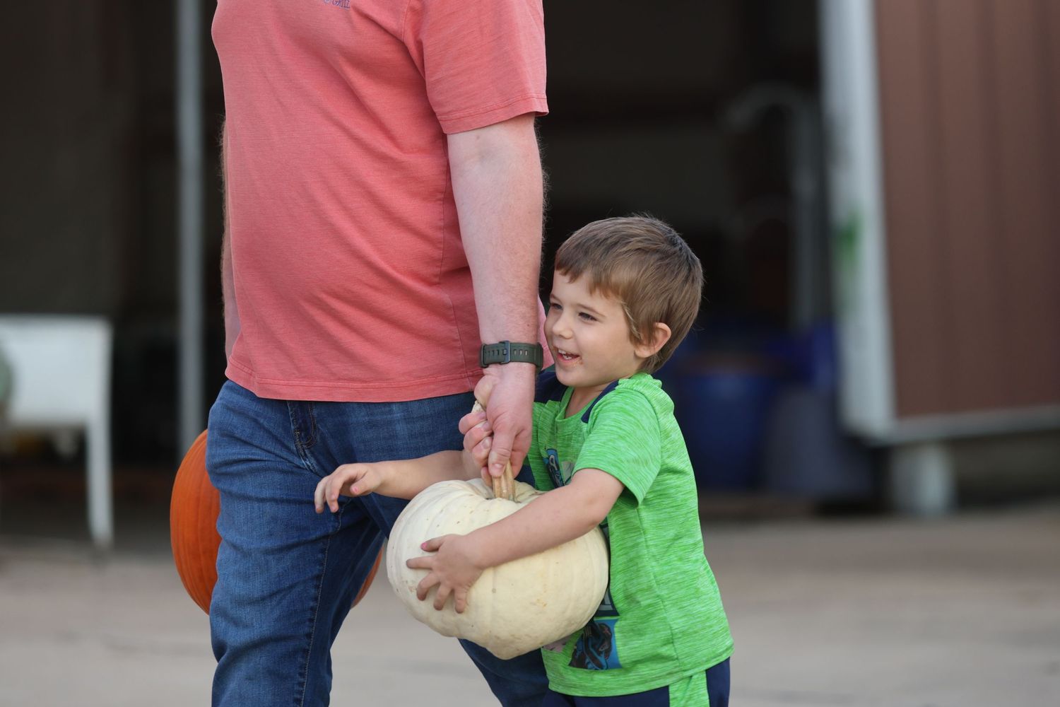 Young child in bright green shirt carries a pumpkin next to an adult in a pink shirt.