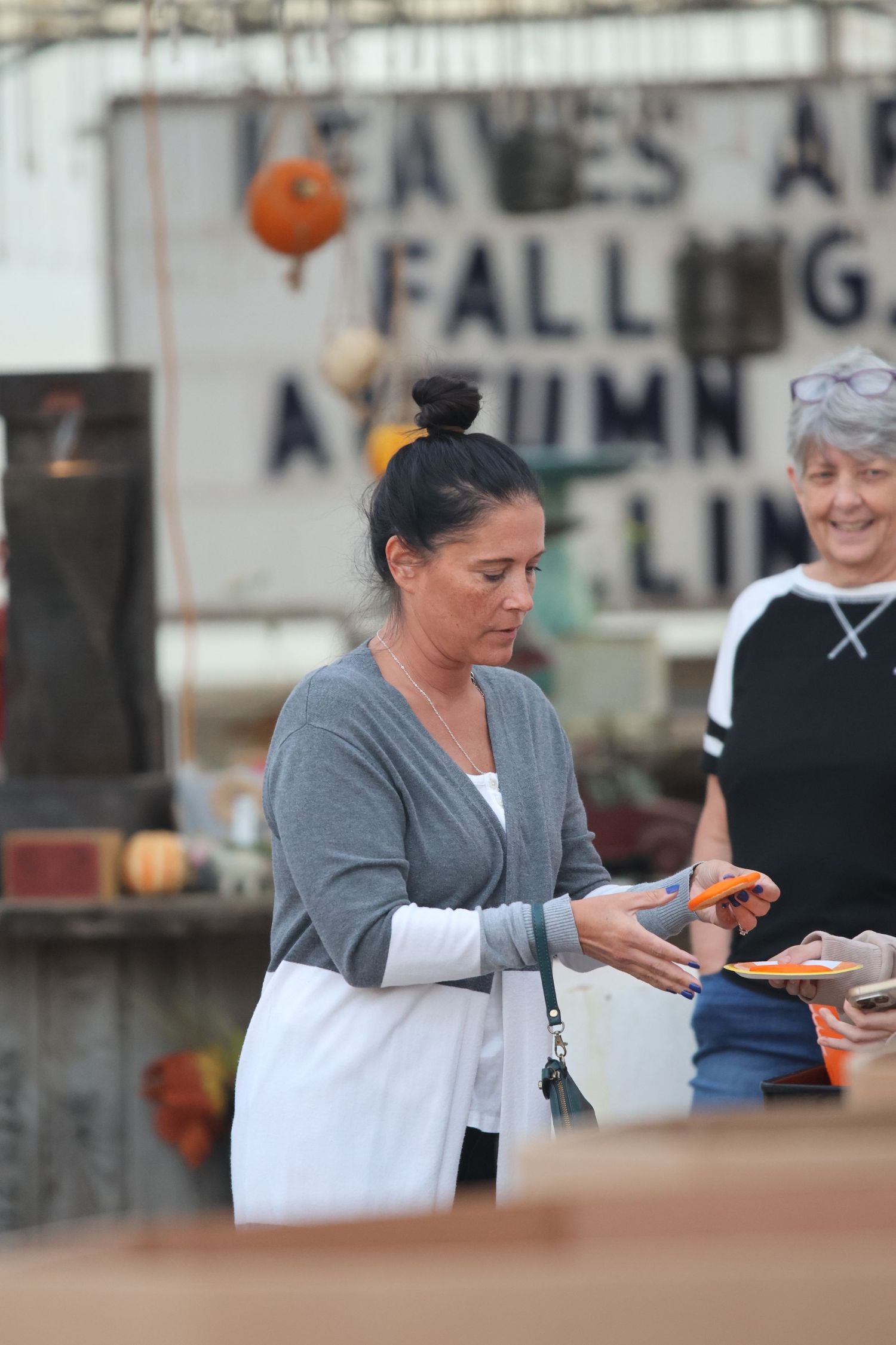 Woman wearing a gray cardigan and white dress receives food at an outdoor event with autumn decorations in the background.