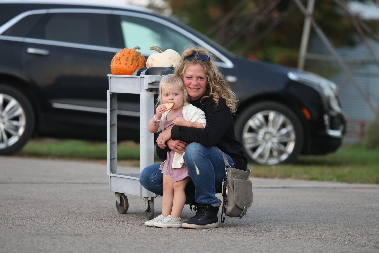 Person sitting with child on rolling cart in parking lot with pumpkins.
