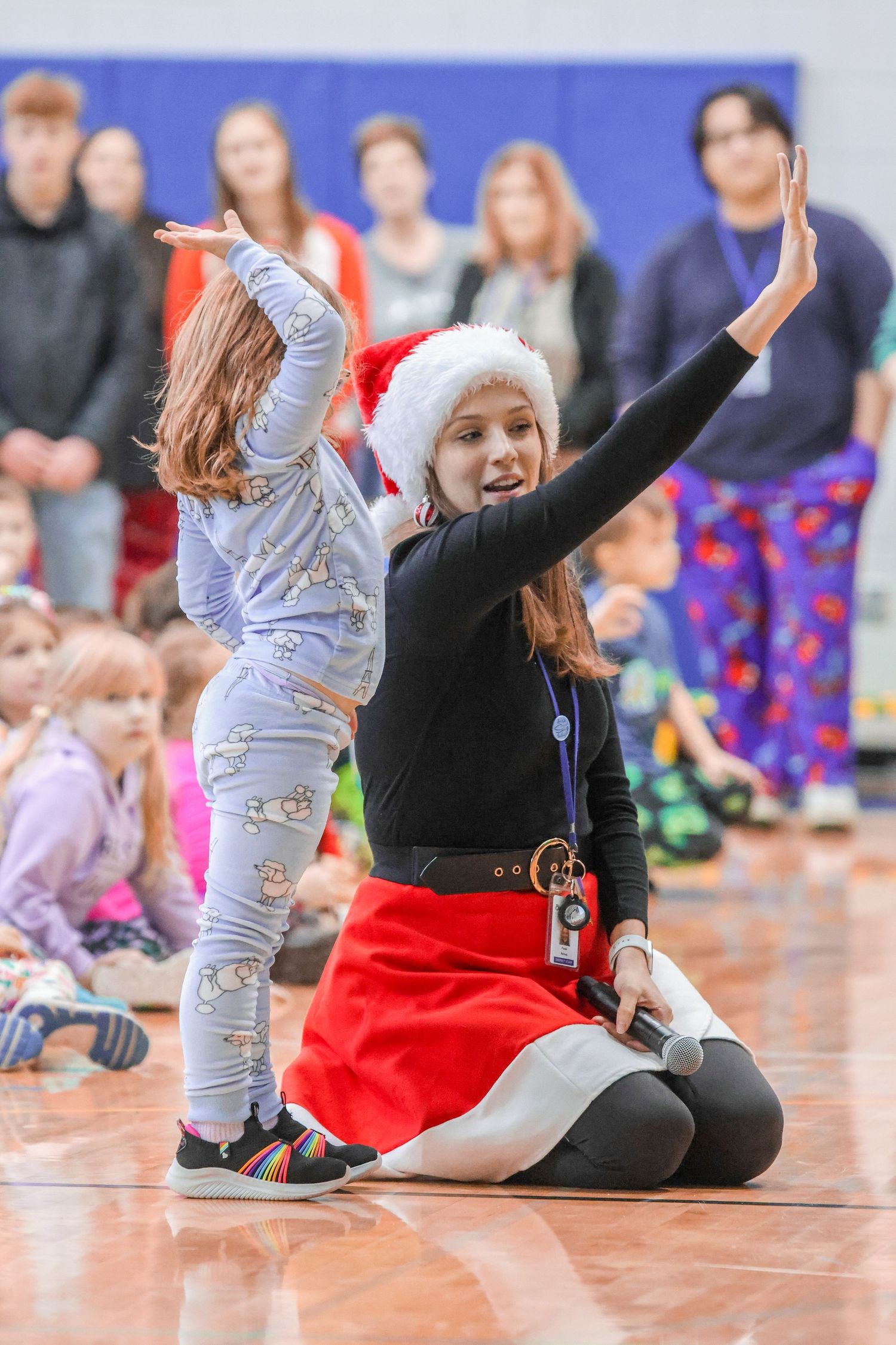 Music director in Santa hat and red skirt leads choir ensamble with young child in gymnasium.