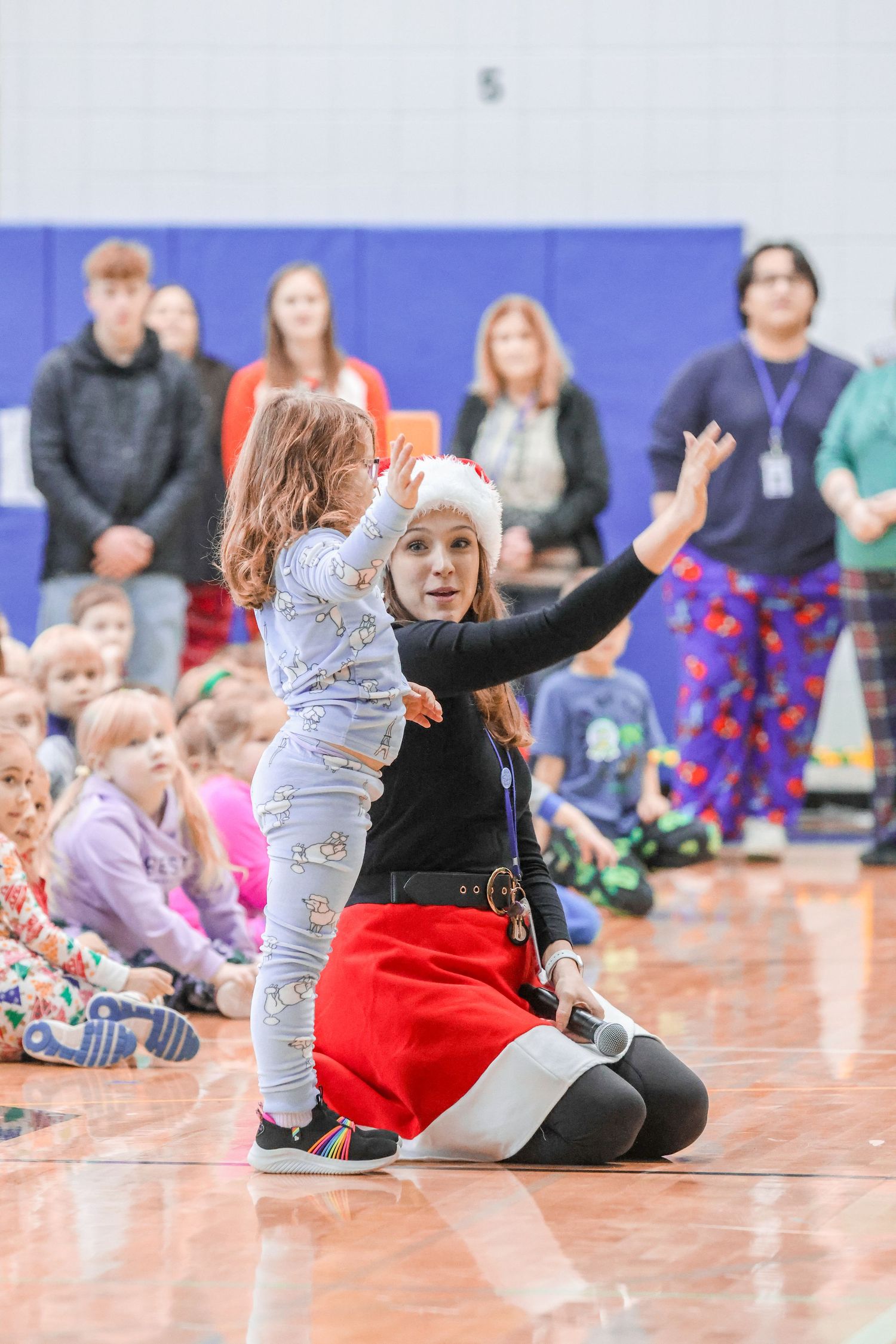 Musical activity in gymnasium with instructor in red skirt and child in pajamas.