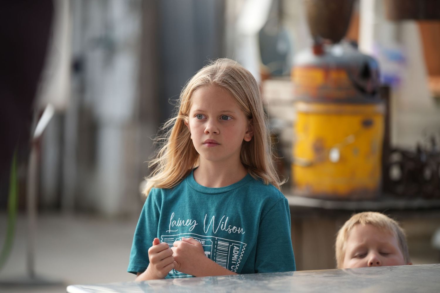 Child in teal t-shirt stands at table in casual indoor setting.