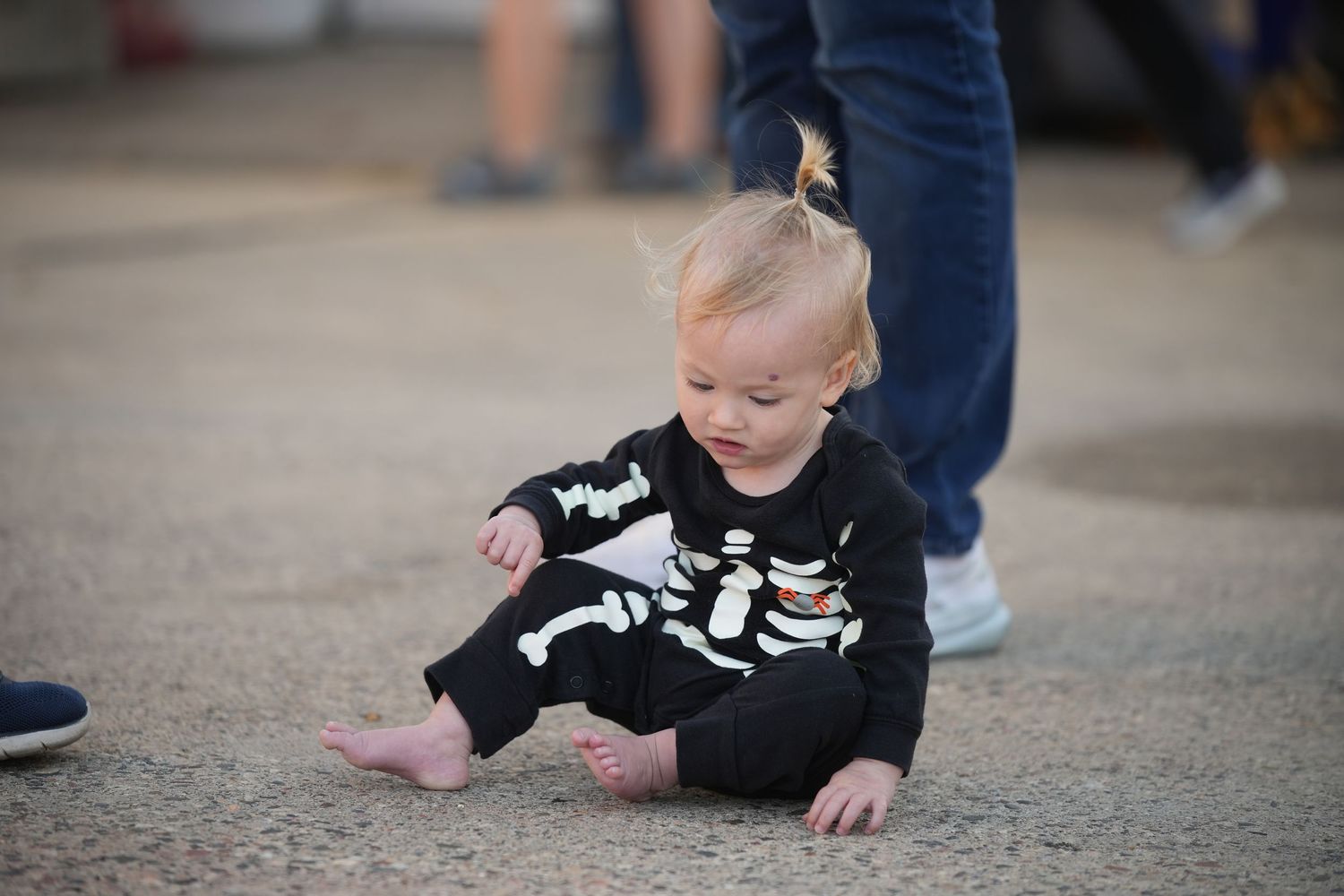 Baby wearing black skeleton outfit sits on carpet floor with pink socks.