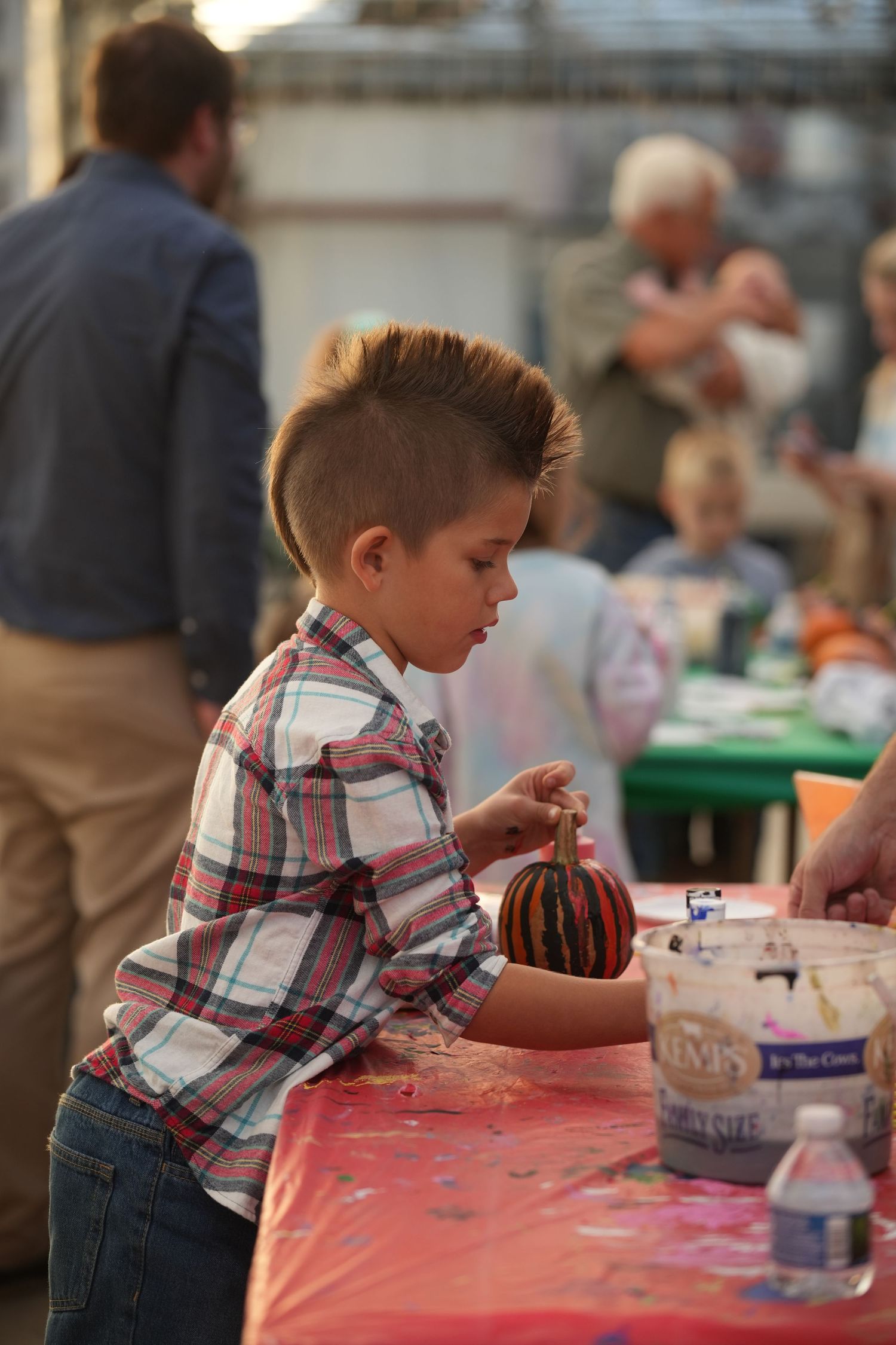 Young child in plaid shirt sits at craft table working intently on an art project at community event.