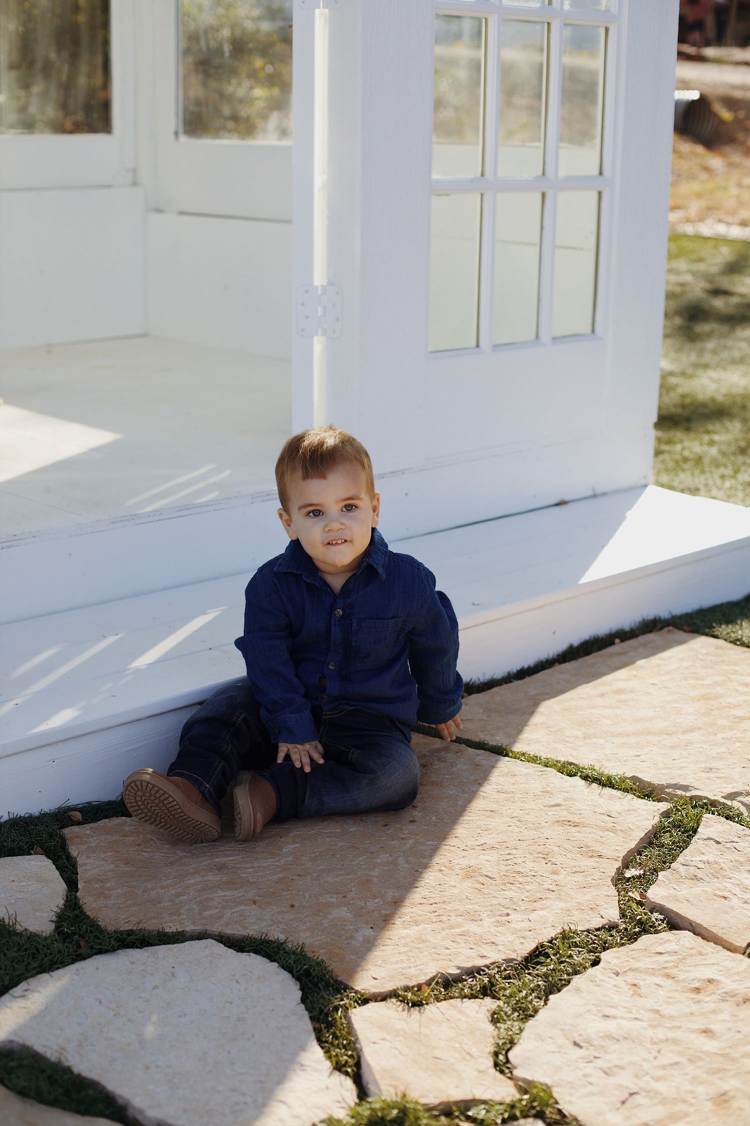 Portrait of little boy sitting on the ground near a glasshouse.