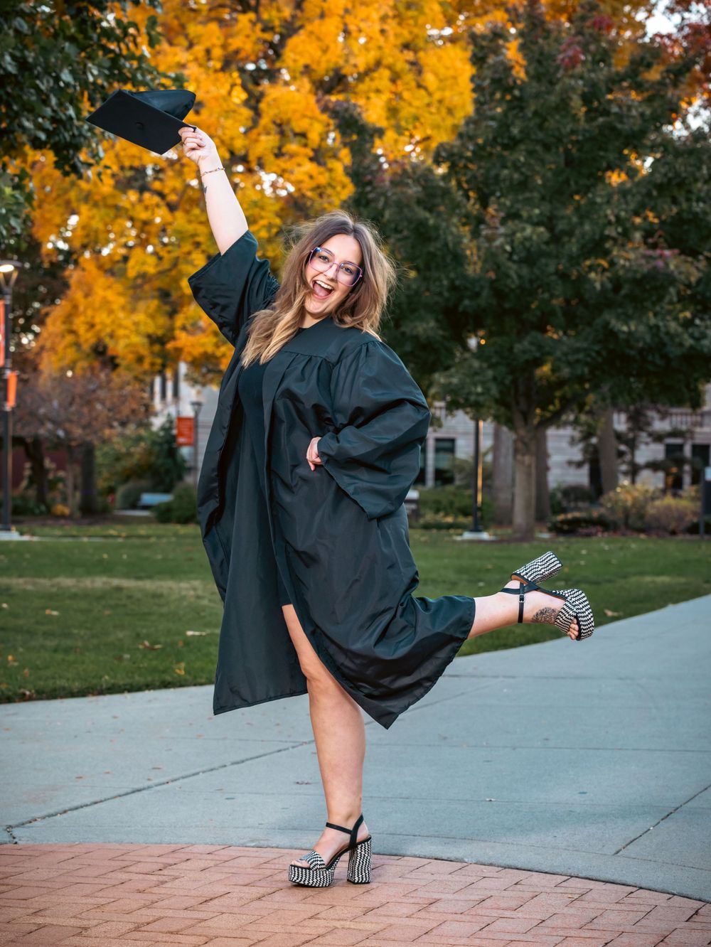 A graduate in black gown strikes a playful pose with autumn foliage in the background.