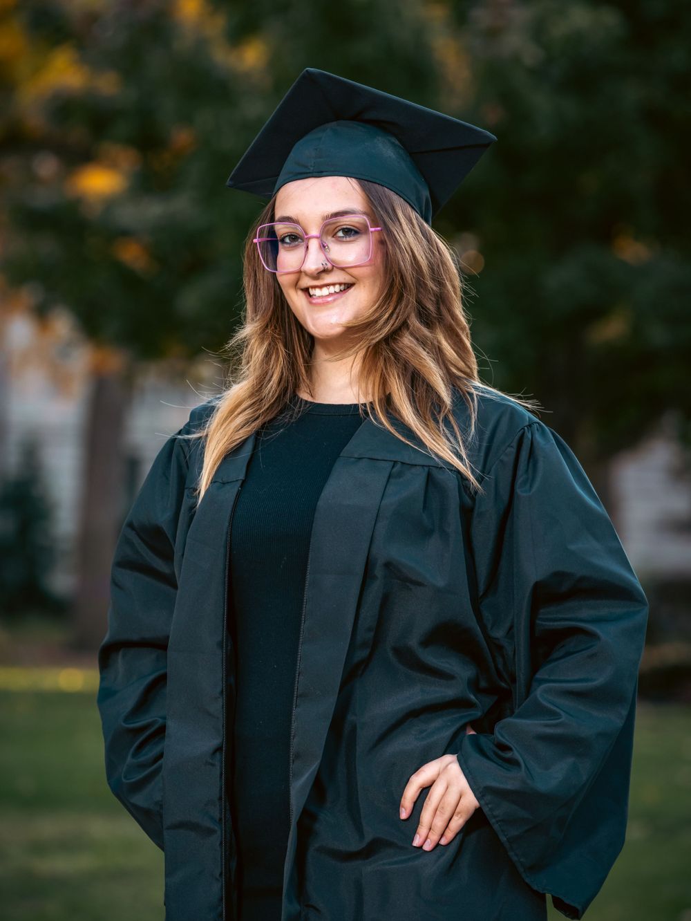 A graduate in cap and gown poses outdoors during autumn.
