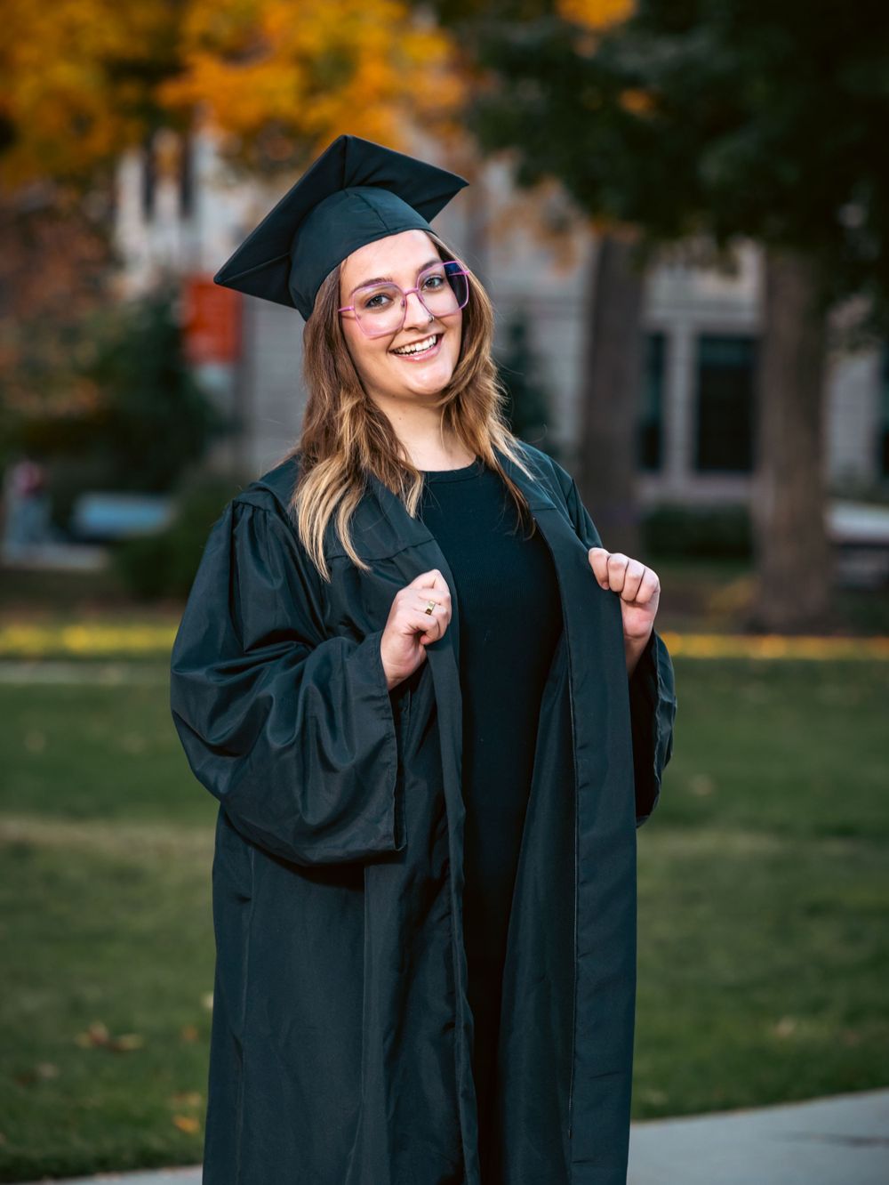 A graduate in academic dress smiles while posing on campus grounds.