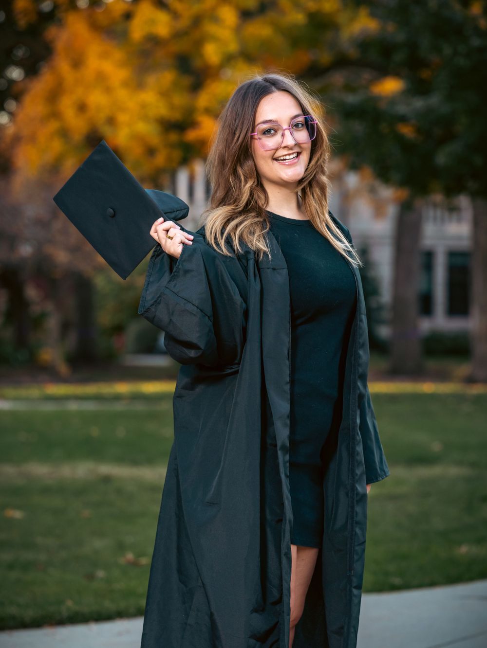 A graduate holds their diploma while posing outdoors in academic regalia.