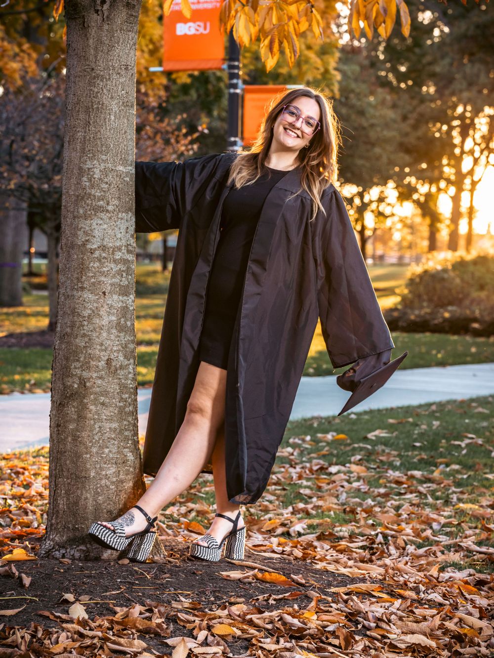Graduate posing in cap and gown by tree during golden sunset with autumn leaves and sparkly shoes.