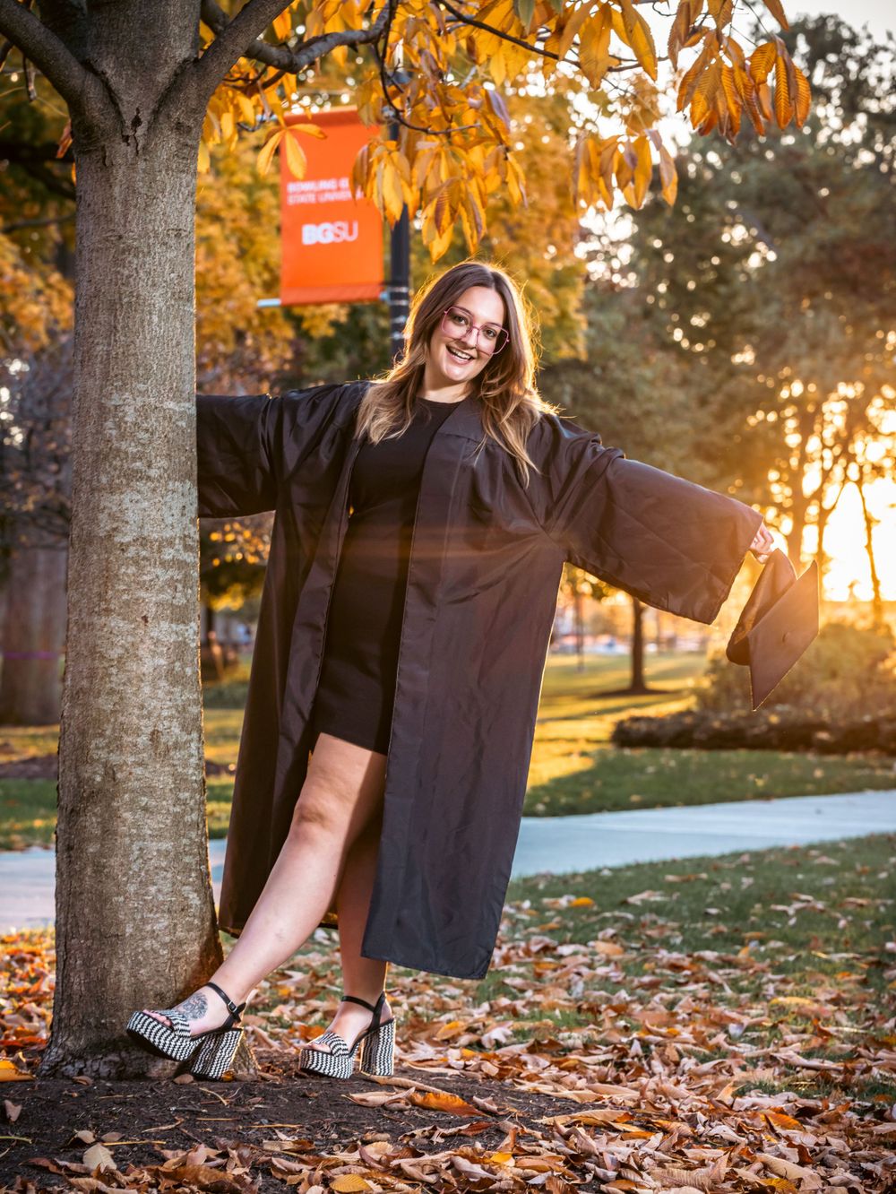 Playful graduation photo showing graduate leaning against tree at sunset in sparkling heels.
