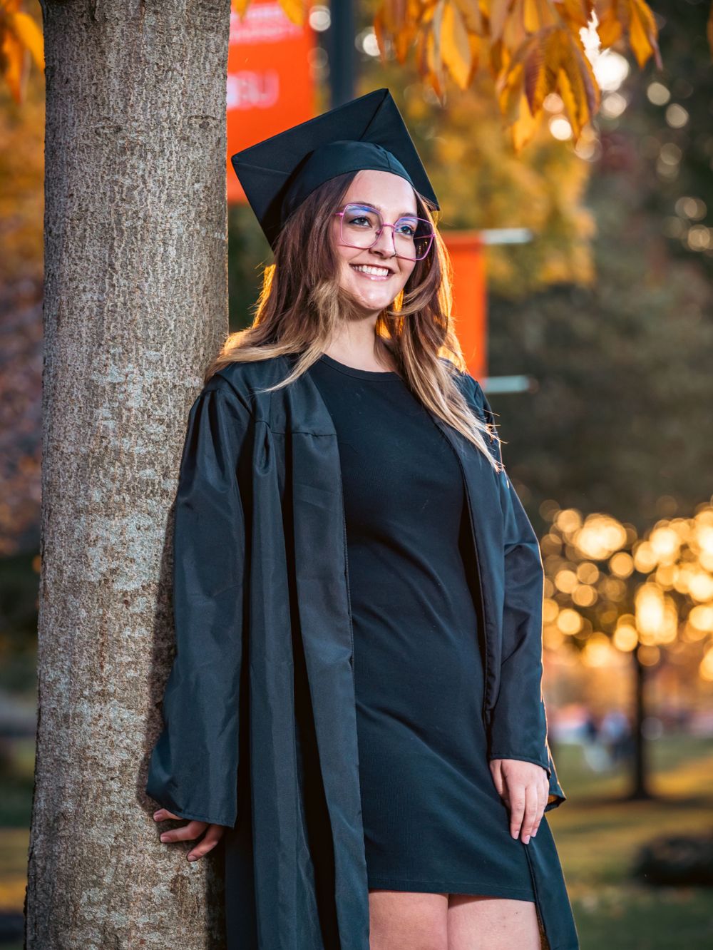 Graduate in traditional black gown and cap smiling by tree with autumn foliage at sunset.