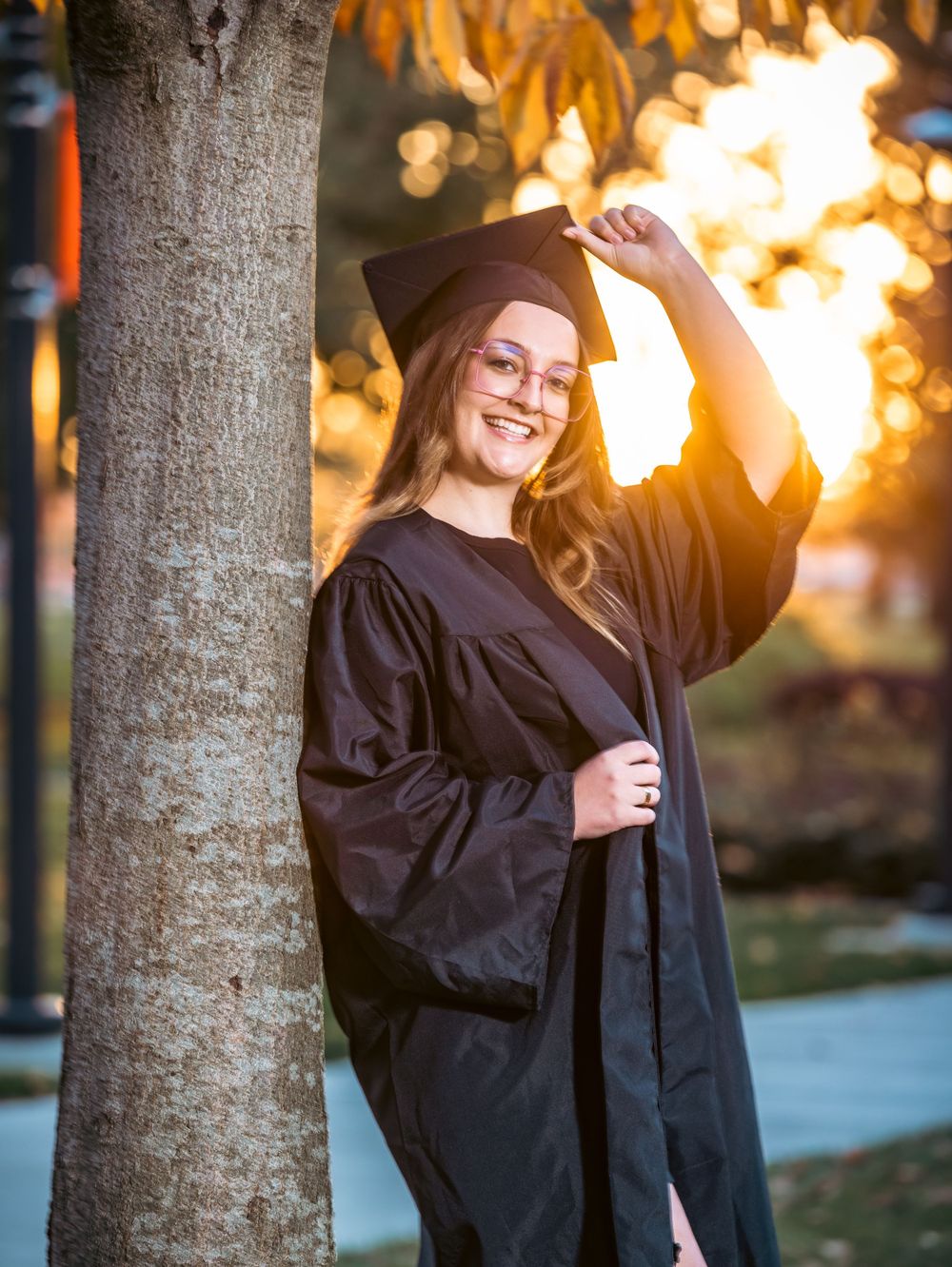 Graduate adjusting mortarboard cap while posing by tree during sunset photoshoot.