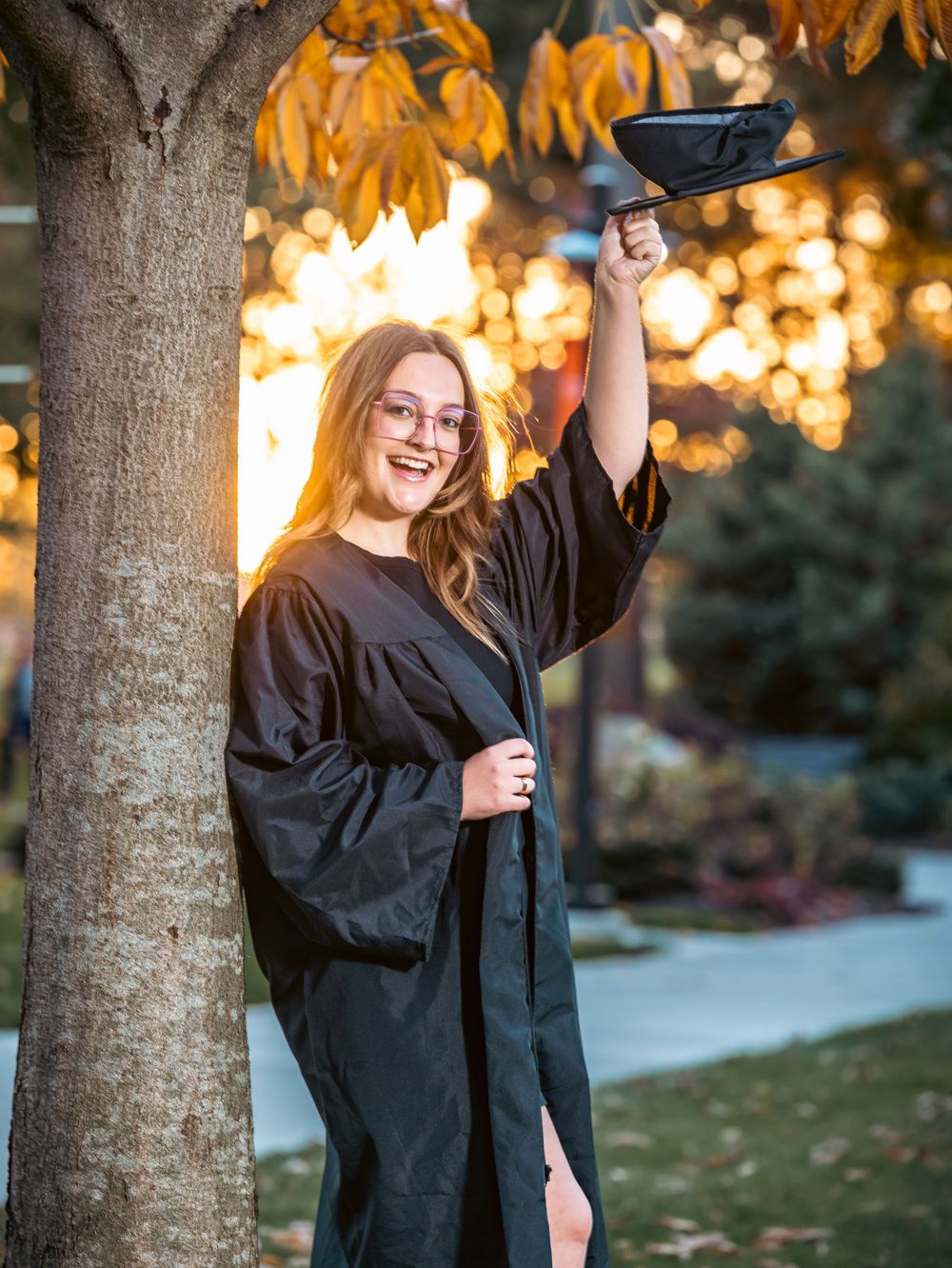 Excited graduate celebrating with diploma held high under autumn tree at golden hour.