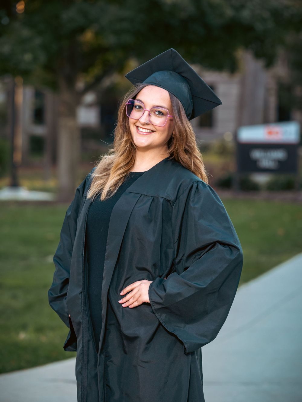 A graduate poses in traditional black cap and gown on a campus pathway.