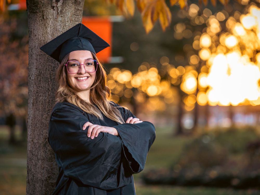 Graduate in academic regalia poses confidently against tree with sunset bokeh background.