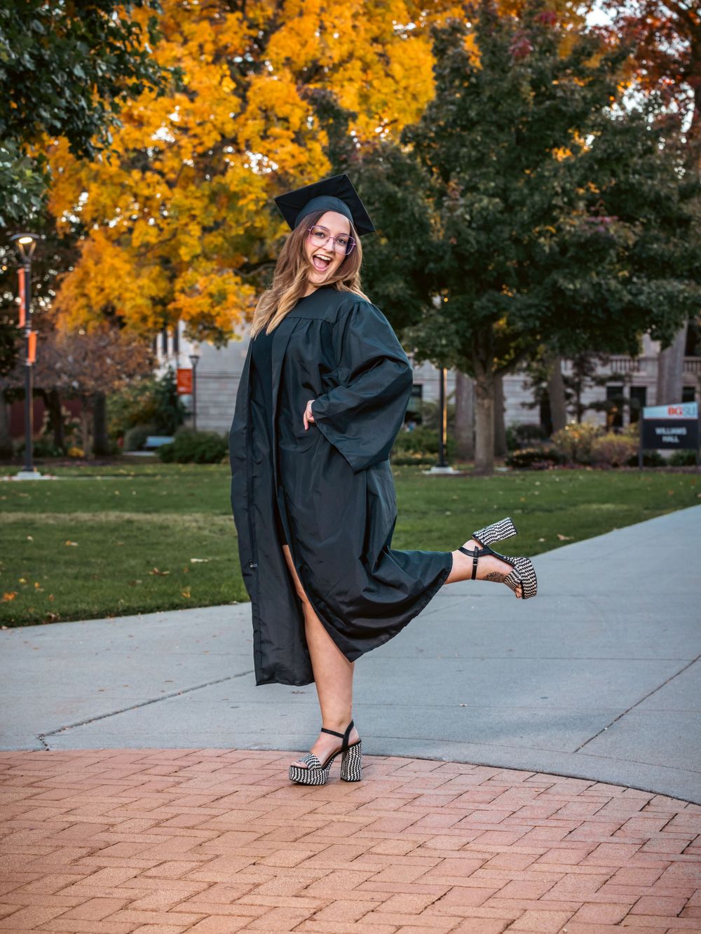 A graduate in academic regalia poses on a brick pathway with fall colors behind.