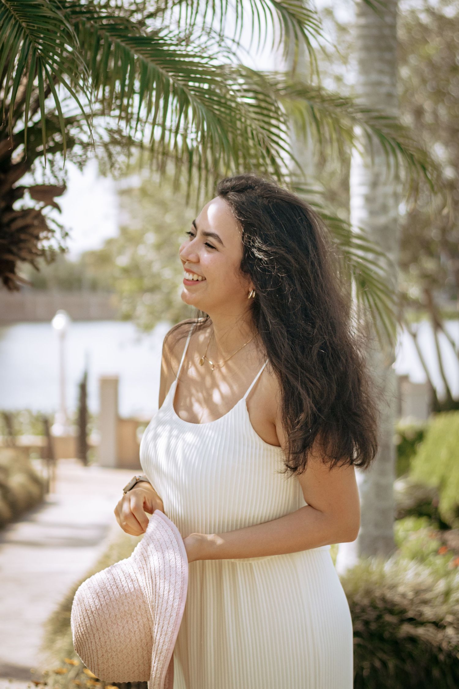 Young woman in white ribbed maxi dress holding a pink hat smiling near tropical palm trees.