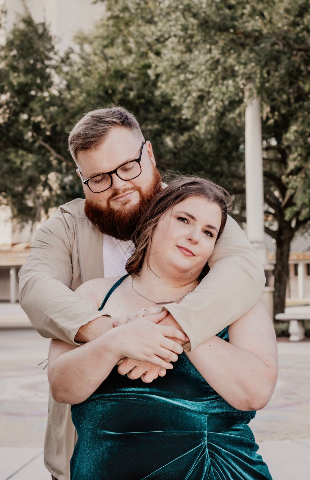 Loving couple embracing outdoors, man with beard hugging woman in green velvet dress near trees.