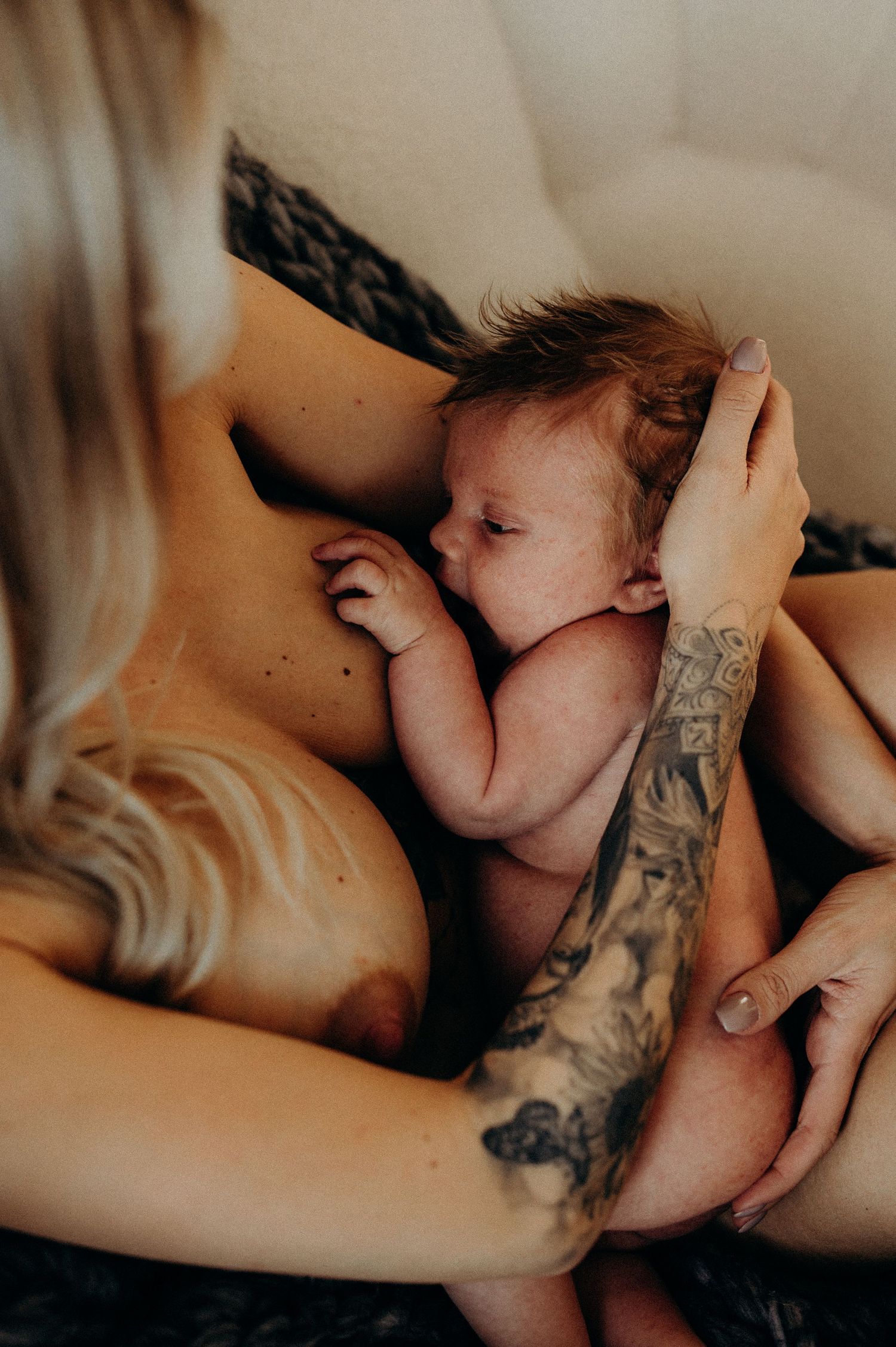 nyföddfotografering i hemmet i stockholm, varm och lite mörkare hudnära bild på son som ligger i mammas famn och ammar.