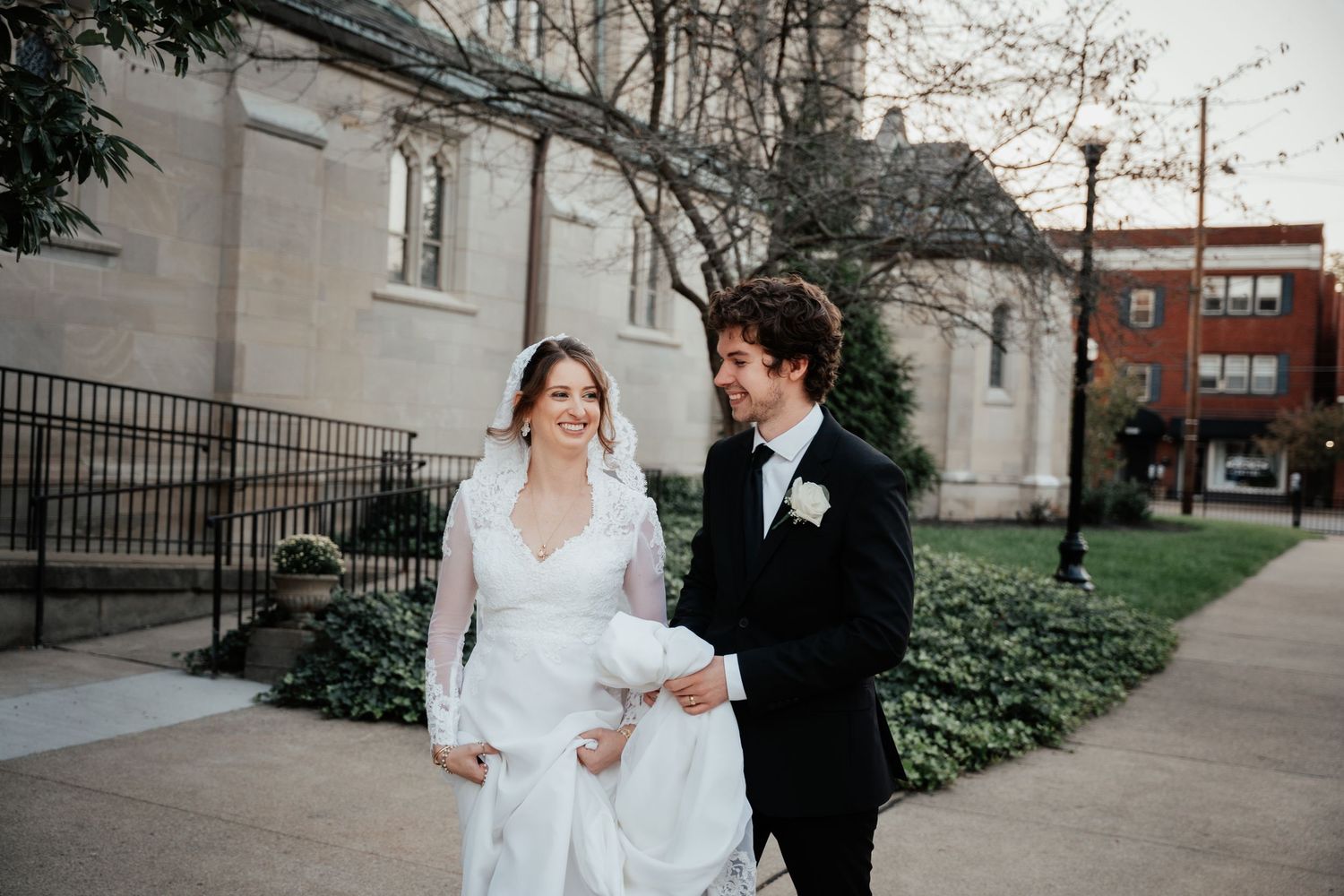 A bride and groom walking together on a pathway outside a church with white stone walls and landscaped grounds.