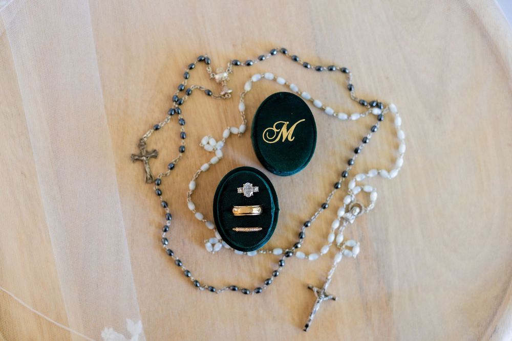 Wedding rings displayed in dark green velvet boxes with a rosary bead necklace arranged on white fabric.