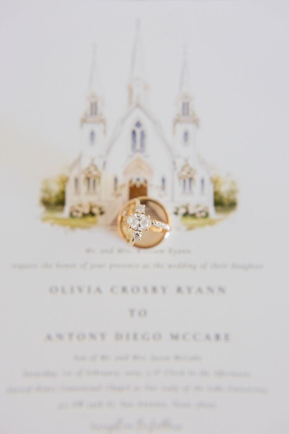 Wedding ring photographed against elegant wedding invitation with white church steeples in background.