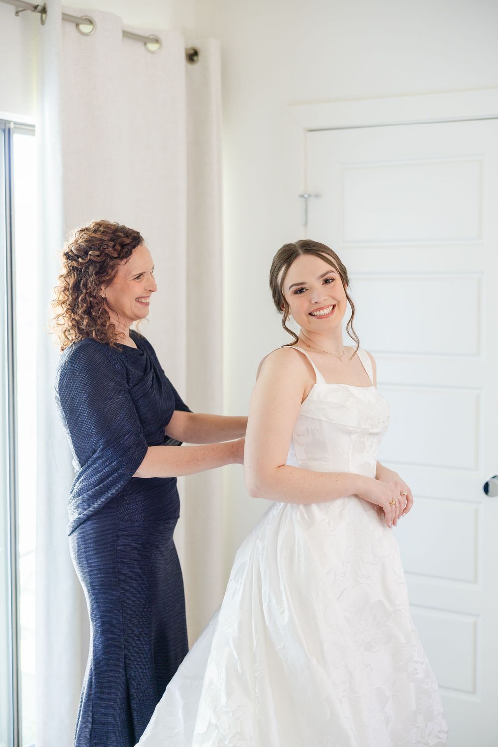 Someone in a navy blue dress helps adjust a white wedding gown in a bright room with sheer curtains.