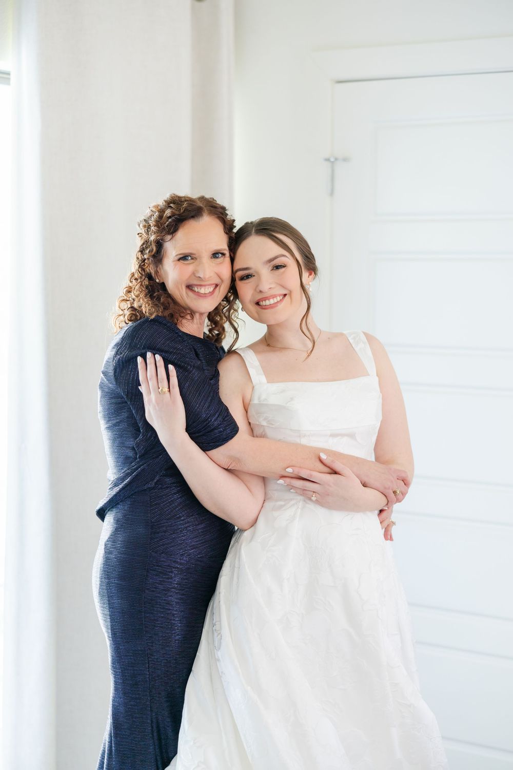Two people embrace and smile while wearing a navy dress and white wedding gown against a light wall.